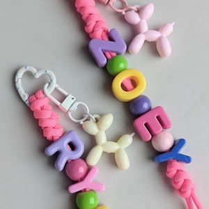 May include: Two keychains with pink braided cords and colorful beads spelling out names. One keychain has a heart-shaped clasp and a pink balloon dog charm. The other has a white heart-shaped clasp and a white balloon dog charm.