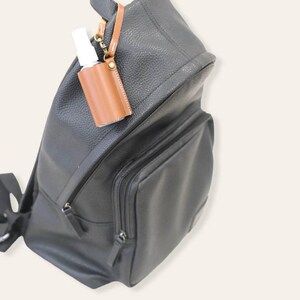 K&ouml;nnte beinhalten: Ein schwarzer Lederrucksack mit einem braunen Lederhalter f&uuml;r Handdesinfektionsmittel, der an der Vorderseite befestigt ist. Der Halter hat einen Metallclip und eine kleine durchsichtige Flasche mit Handdesinfektionsmittel im Inneren.