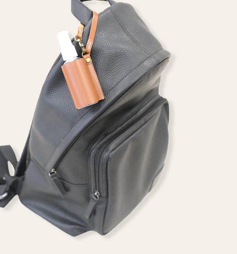K&ouml;nnte beinhalten: Ein schwarzer Lederrucksack mit einem braunen Lederhalter f&uuml;r Handdesinfektionsmittel, der an der Vorderseite befestigt ist. Der Halter hat einen Metallclip und eine kleine durchsichtige Flasche mit Handdesinfektionsmittel im Inneren.