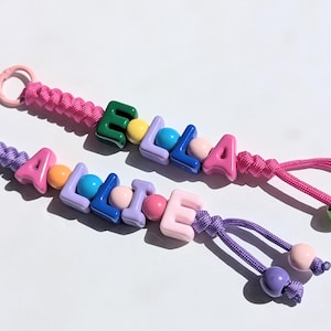 May include: Two handmade keychains spelling out "ALICE" and "ELLA" with colorful letter beads. The keychains feature pink and purple braided cords, with matching beads and loops for attaching keys or accessories. The letters are in various colors.