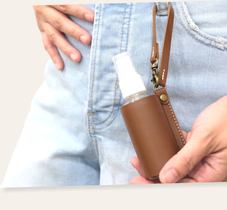 K&ouml;nnte beinhalten: Ein brauner Leder-Handdesinfektionsmittelhalter mit einer durchsichtigen Plastikflasche und einem silbernen Metallverschluss. Der Halter hat einen Riemen zum Befestigen an einer G&uuml;rtelschlaufe oder einer Tasche.