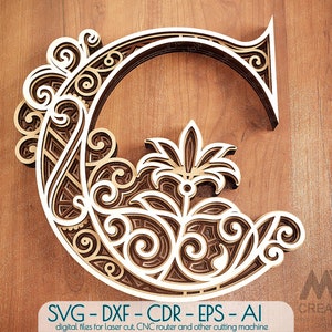 Pode incluir: Um design intrincado de corte a laser da letra G com um padrão floral. O design é em camadas e tem um efeito 3D. A imagem está sobre um fundo de madeira.