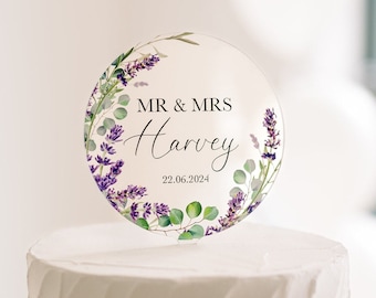 May include: A white wedding cake with textured frosting, topped with a clear, circular cake topper. The topper features the text "MR & MRS Harvey" and the date "22.06.2024", surrounded by a floral design with purple lavender and green leaves.
