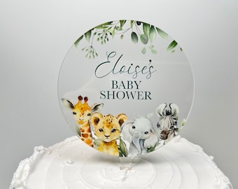 May include: A clear, round cake topper with a watercolor design featuring a giraffe, lion, elephant, and zebra. The text "Eloise's BABY SHOWER" is in green script. Green leaves and foliage frame the top.