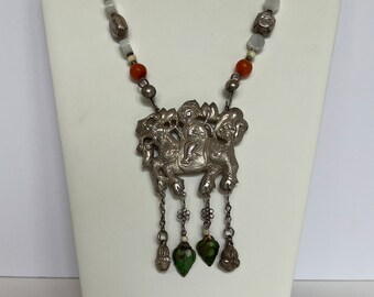 May include: Silver-toned necklace with a detailed pendant featuring a mythical creature design. The necklace includes orange, white, and gray beads, with green teardrop-shaped accents hanging from the pendant. The necklace is displayed on a white mannequin.