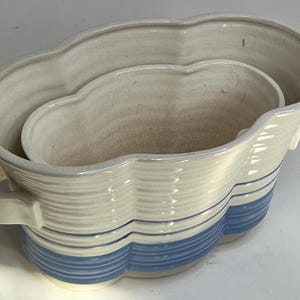 May include: A ceramic planter with a wavy design, featuring a cream-colored exterior with blue stripes and a matching interior. The planter has a handle on one side and a nested interior section.