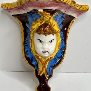 May include: A decorative wall sconce with a painted face. The sconce features a white face with a stern expression, framed by yellow and blue accents. The top is yellow with red and brown details.