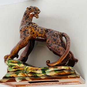 May include: A ceramic figurine of a leopard with brown and black spots, standing on a green, brown, and white base. The leopard is in a leaping pose with its tail curled around its body.