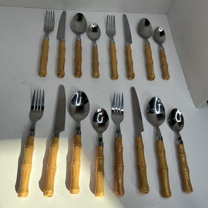 May include: A set of stainless steel silverware with bamboo-style handles. The set includes forks, knives, and spoons in various sizes. The bamboo handles are a light brown color, and the metal parts are shiny.