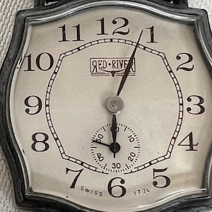 May include: A vintage Red River wristwatch with a cream-colored face and black numerals. The watch features a small seconds dial and a brown leather strap. The watch face has the text "RED RIVER" and "SWISS".