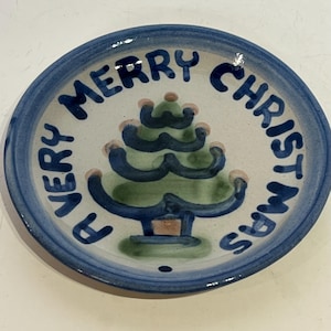 May include: A small, round ceramic plate with a blue rim and a white interior. The plate features a hand-painted Christmas tree in the center, surrounded by the words "VERY MERRY CHRISTMAS" in blue lettering.