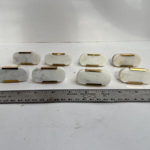 May include: Eight marble and gold cabinet knobs. Each knob is a rectangular shape with rounded edges. The marble is white with gray veining, and the gold is a thin band across the top. A ruler is in the foreground.