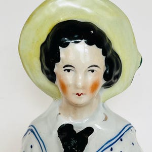 May include: A vintage porcelain doll with a pale yellow brimmed hat and black hair. The doll has a white sailor-style top with blue polka dots and trim. The doll's face has rosy cheeks and red lips.