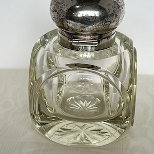May include: A clear glass inkwell with a silver-colored, spherical lid. The inkwell has a square base with rounded corners and a decorative, starburst pattern on the bottom. The lid is hinged and has a tarnished appearance.