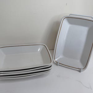 May include: A set of five rectangular white ceramic dishes with rounded corners and a thin brown line accent along the inner rim. One dish is displayed upright on a stand, while the other four are stacked horizontally in a neat pile.