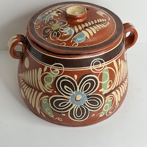 May include: A round, terracotta-colored ceramic pot with a lid, featuring hand-painted floral designs in white, blue, and green. The pot has two handles and a small knob on the lid. The design includes a large flower on the front.