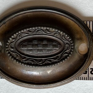 May include: An oval-shaped, antique brass drawer pull with a decorative, woven design in the center. The pull has two holes for mounting.