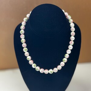 May include: A white beaded necklace with pink and green floral designs. The necklace is displayed on a black mannequin. The beads are round and evenly spaced, creating a delicate and feminine accessory.