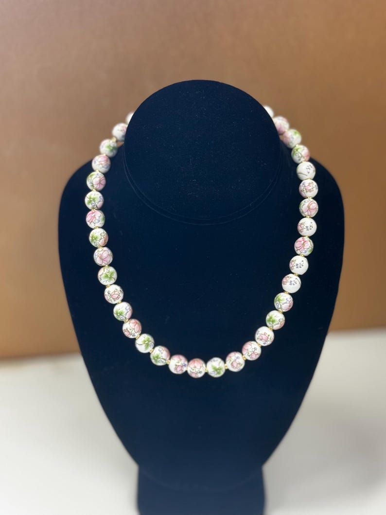 May include: A white beaded necklace with pink and green floral designs. The necklace is displayed on a black mannequin. The beads are round and evenly spaced, creating a delicate and feminine accessory.
