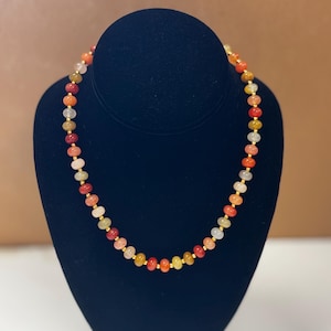 May include: A colorful beaded necklace displayed on a black velvet bust. The necklace features a variety of small, round beads in shades of red, orange, yellow, and white, strung together with gold-colored spacers. The necklace is a jewelry item.