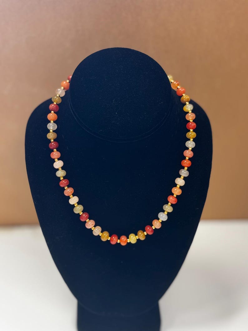 May include: A colorful beaded necklace displayed on a black velvet bust. The necklace features a variety of small, round beads in shades of red, orange, yellow, and white, strung together with gold-colored spacers. The necklace is a jewelry item.