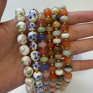 May include: Several beaded necklaces in various styles and colors. Some necklaces feature round, painted ceramic beads with floral and blue patterns. Others have colorful gemstone beads in shades of orange, green, and blue, strung with gold-colored spacers.