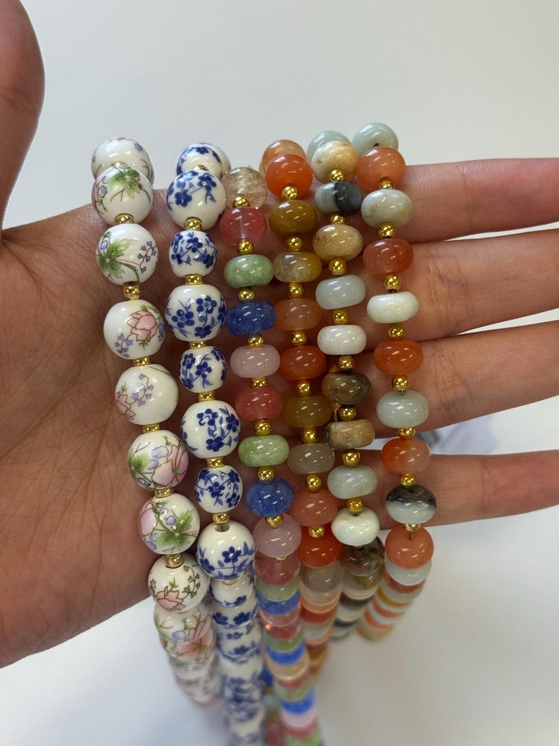 May include: Several beaded necklaces in various styles and colors. Some necklaces feature round, painted ceramic beads with floral and blue patterns. Others have colorful gemstone beads in shades of orange, green, and blue, strung with gold-colored spacers.