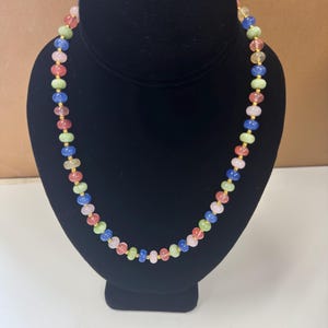 May include: A colorful beaded necklace displayed on a black bust. The necklace features a variety of small, round beads in shades of pink, green, blue, and yellow, strung together with gold-colored chain links. The necklace is a vibrant accessory.
