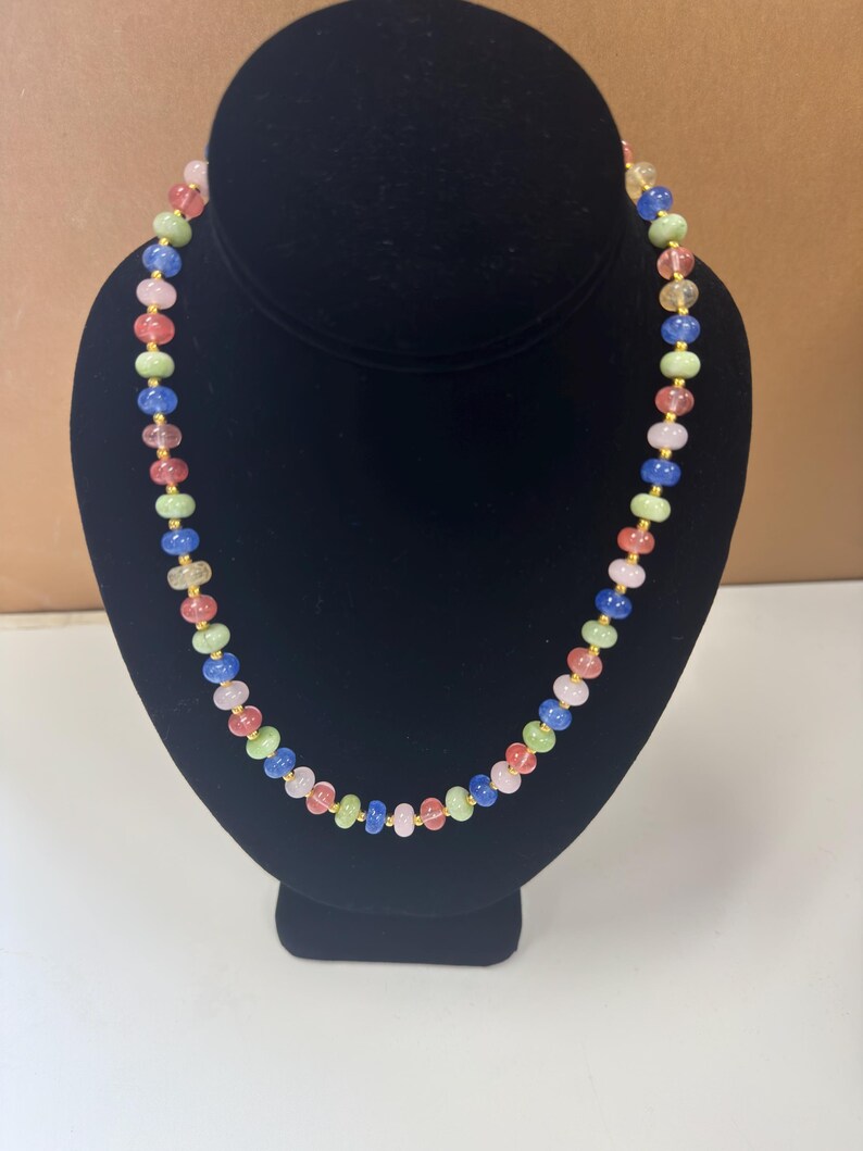 May include: A colorful beaded necklace displayed on a black bust. The necklace features a variety of small, round beads in shades of pink, green, blue, and yellow, strung together with gold-colored chain links. The necklace is a vibrant accessory.