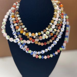 May include: A display of beaded necklaces in various colors, including white, blue, red, orange, and green. The necklaces are arranged on a black mannequin. Some beads have floral patterns, and others are solid.
