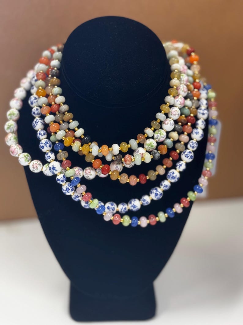 May include: A display of beaded necklaces in various colors, including white, blue, red, orange, and green. The necklaces are arranged on a black mannequin. Some beads have floral patterns, and others are solid.