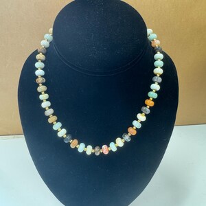May include: A beaded necklace with a variety of colorful, irregular-shaped stones. The stones are in shades of green, orange, brown, and cream, interspersed with small gold-colored beads. The necklace is displayed on a black bust.