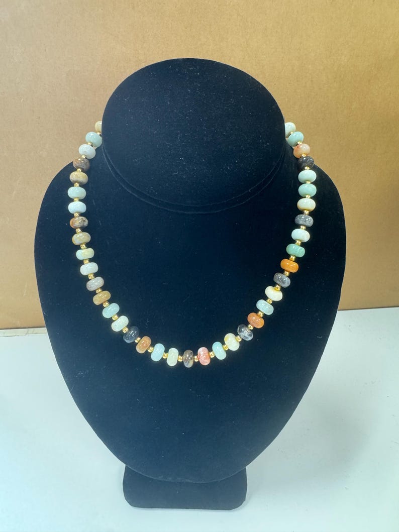 May include: A beaded necklace with a variety of colorful, irregular-shaped stones. The stones are in shades of green, orange, brown, and cream, interspersed with small gold-colored beads. The necklace is displayed on a black bust.