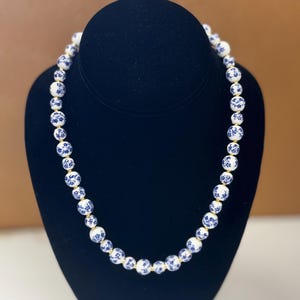 May include: A beaded necklace featuring white and blue porcelain beads. The beads are adorned with a blue floral pattern and strung together with a gold-colored cord. The necklace is displayed on a black bust.