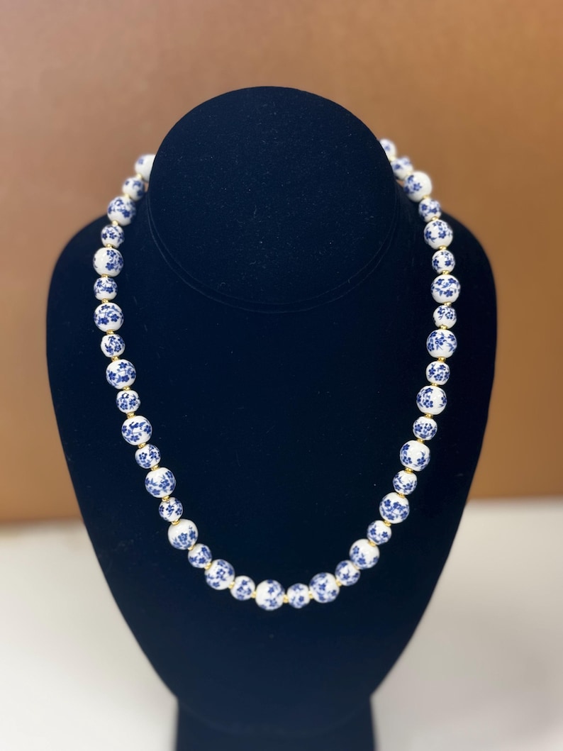 May include: A beaded necklace featuring white and blue porcelain beads. The beads are adorned with a blue floral pattern and strung together with a gold-colored cord. The necklace is displayed on a black bust.