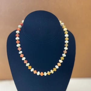 May include: A beaded necklace with a variety of colorful stones, including orange, white, and yellow, interspersed with small gold-colored beads. The necklace is displayed on a black bust.