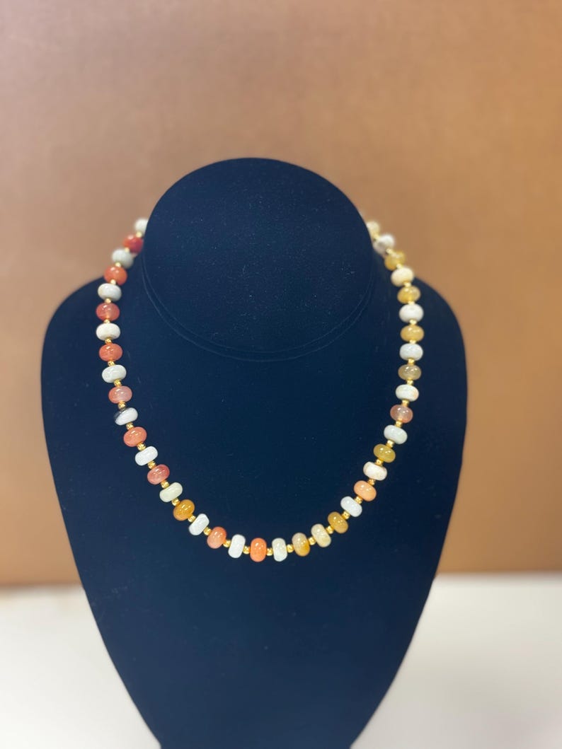 May include: A beaded necklace with a variety of colorful stones, including orange, white, and yellow, interspersed with small gold-colored beads. The necklace is displayed on a black bust.