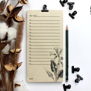 Könnte beinhalten: Ein braunes Papier-Notizblock für To-Do-Listen mit einem floralen Design und dem aufgedruckten Text "TO-DO" und "NOA". Der Notizblock liegt auf einer weißen Oberfläche mit einem grünen Bleistift und schwarzen Büroklammern.