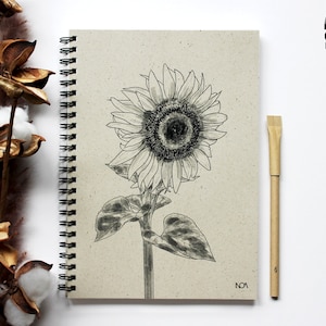 Könnte beinhalten: Ein beigefarbenes Notizbuch mit Spiralbindung, auf dem eine Schwarzweißzeichnung einer Sonnenblume abgebildet ist. Das Notizbuch trägt die Aufschrift "NOA" in der unteren rechten Ecke. Ein Holzstift liegt neben dem Notizbuch.
