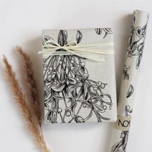 Könnte beinhalten: Ein verpacktes Geschenk und eine Rolle Geschenkpapier mit einem schwarz-weißen Mistelzweig-Design. Das Geschenk ist mit einem natürlichen Bastband gebunden. Geschenkpapier und Schachtel haben einen gesprenkelten, hellgrauen Hintergrund.