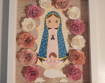May include: A framed illustration of the Virgin Mary with a pink, white, and blue paper flower border. The Virgin Mary is depicted in a blue robe with gold stars and a pink dress. She is holding a baby Jesus in her arms. The illustration is surrounded by a white frame.