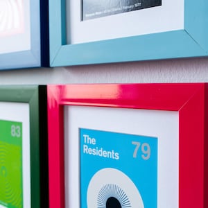 May include: A collection of framed art prints with colorful frames. The frames are in various colors, including red, blue, and green. The prints feature graphic designs and text, including "The Residents 79".