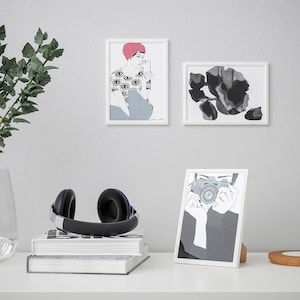 May include: Three white picture frames with black and white illustrations. The top left frame shows a woman with a pink hat and a shirt with eye designs. The top right frame shows a black and white abstract design. The bottom frame shows a person holding a camera.