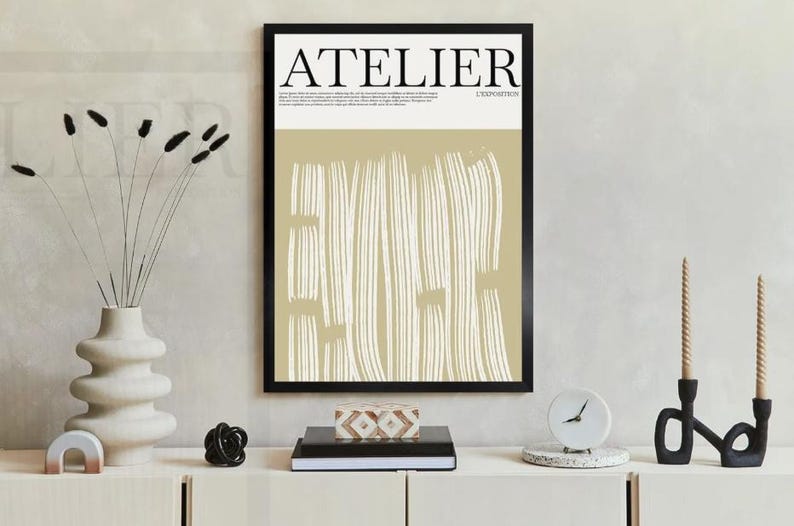 May include: A framed art print with the word "ATELIER" in black letters. The artwork features a beige background with vertical white brushstrokes. A white vase with black stems and a decorative clock are on a white cabinet.