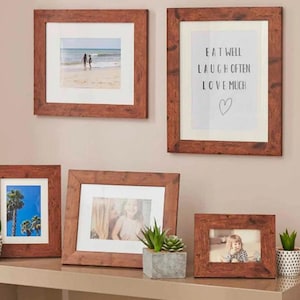 Puede incluir: Una colección de cuatro marcos de fotos de madera marrón con diferentes imágenes en su interior. Los marcos están colgados en una pared y hay pequeñas plantas en macetas en un estante debajo de los marcos. El texto en uno de los marcos dice "Come bien, ríe a menudo, ama mucho".