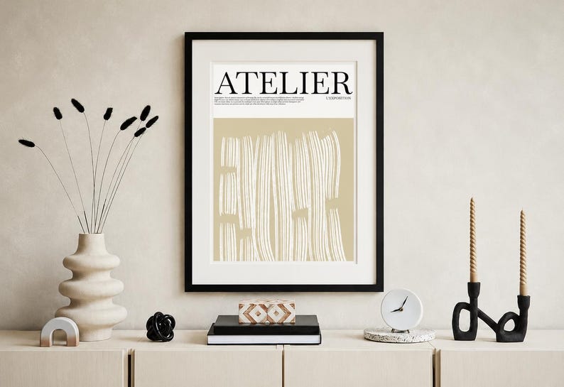 May include: A framed art print with the word "ATELIER" in black letters. The print has a beige background with white vertical lines. A vase with dried flowers, a clock, and candles are on a wooden surface.