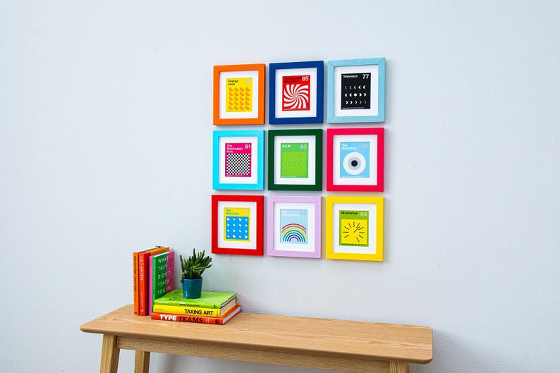 May include: Nine framed art prints with colorful frames are arranged on a white wall. The prints feature various graphic designs in bright colors. A wooden table below holds books and a small potted plant.
