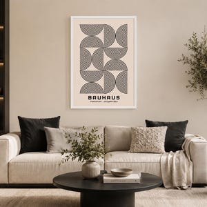 May include: A framed Bauhaus art print with a cream background and black geometric design hangs above a beige sofa. The print features the word "Bauhaus" and the text "Frankfurt - Oktober 1923". The living room is decorated with neutral tones.