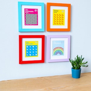 May include: Four framed art prints with colorful frames hang on a white wall. The prints feature text and graphics, including a checkerboard, orange slices, raindrops, and a rainbow. A small potted plant sits on a wooden shelf below.