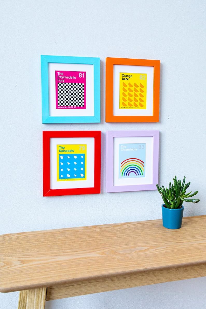 May include: Four framed art prints with colorful frames hang on a white wall. The prints feature text and graphics, including a checkerboard, orange slices, raindrops, and a rainbow. A small potted plant sits on a wooden shelf below.